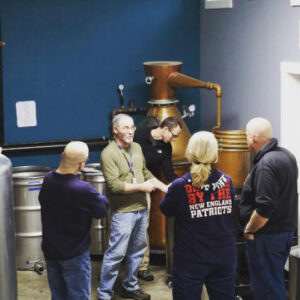 a group of people standing in the distillery being taken around on a tour, listening to a man in a green shirt talking