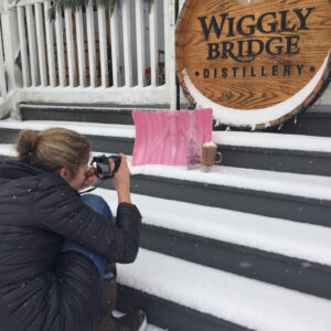 an image of a woman taking a photo outside in the snow of a shovel with a spirit bottle next to a glass